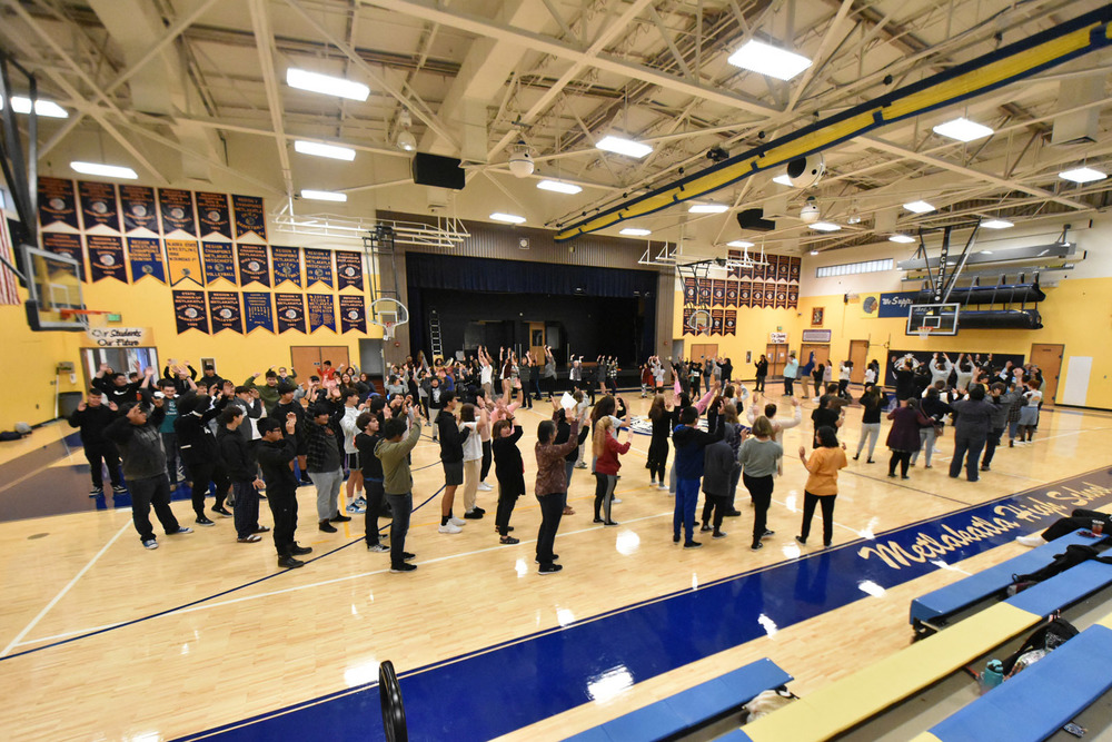 students and staff in gym standing in large circle with their arms up