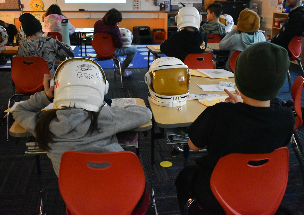 students wearing space helmets, one sits on  a desk