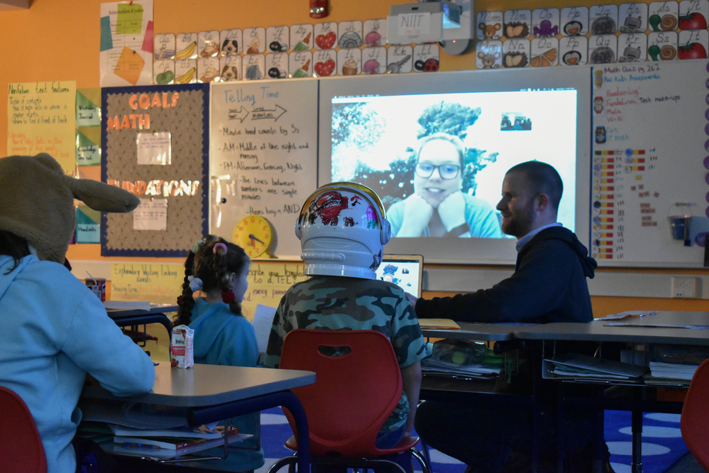 students at desks with a space helmet, woman on projector screen speaking to them