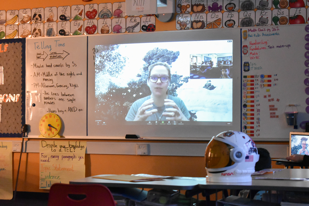 a woman on the screen speaking to the students with a space helmet on the desk